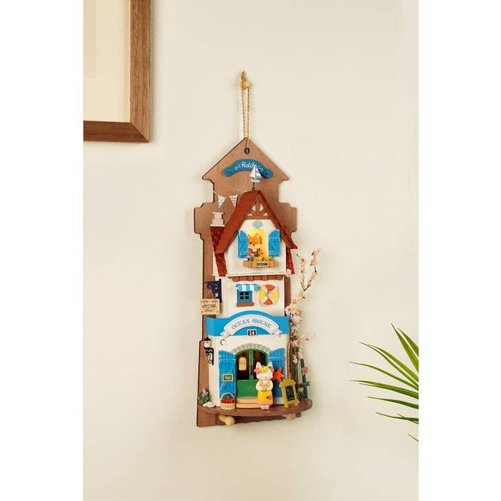 The "Island Dream Villa," a DIY Miniature Wall Hanging Kit from Hands Craft, features a delightful bakery storefront design complete with a blue door, awnings, and charming floral details. It includes a small plant to enhance its appeal, making this piece an ideal gift for both crafters and collectors.