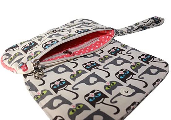 The Bungalow 360 Large Wristlet Cat Print is a white zippered fabric pouch made from natural cotton canvas with a wrist strap. It features an eye-catching pattern of stylized cats with green and blue eyes, while the interior is lined with pink fabric adorned with white polka dots. A small silver charm decorates the zipper pull.