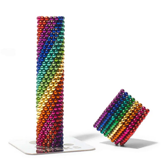 The 512 Rainbow Spectrum Speks by SPEKS are colorful magnetic balls stacked to form two geometric shapes: a tall cylinder and a smaller hexagonal column. The balls are arranged in rainbow order, creating a visually striking and playful pattern.