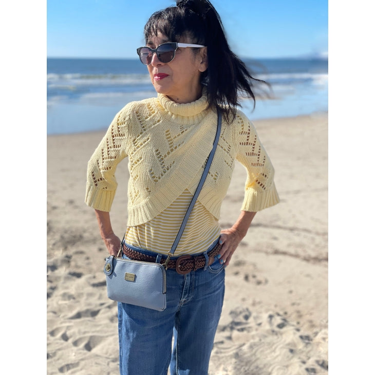 A woman stands on a beach with the ocean in the background, sporting sunglasses, a yellow sweater with a chevron pattern, blue jeans, and a brown belt. She carries the versatile Tina Cell Bag – Black by Liz Soto Handbags in crossbody style and appears to be enjoying a sunny day.