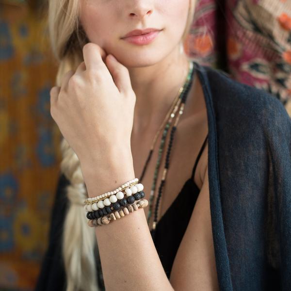 A woman with a relaxed expression is shown from the shoulders up. She has long, blonde hair styled in a loose braid over her shoulder and is wearing a black top with a dark, sheer cover-up. She is accessorized with the Aqua Terra Stone Stacking Bracelet by Scout Jewelry and layered necklaces.
