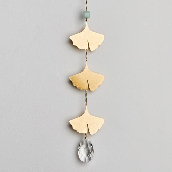 The Leaf Amazonite Suncatcher by Scout Jewelry showcases three gold ginkgo leaf pendants elegantly aligned in a vertical design. It is embellished with a lustrous blue bead at the top and finishes with a teardrop-shaped crystal at the bottom, casting rainbows against a simple light gray background.