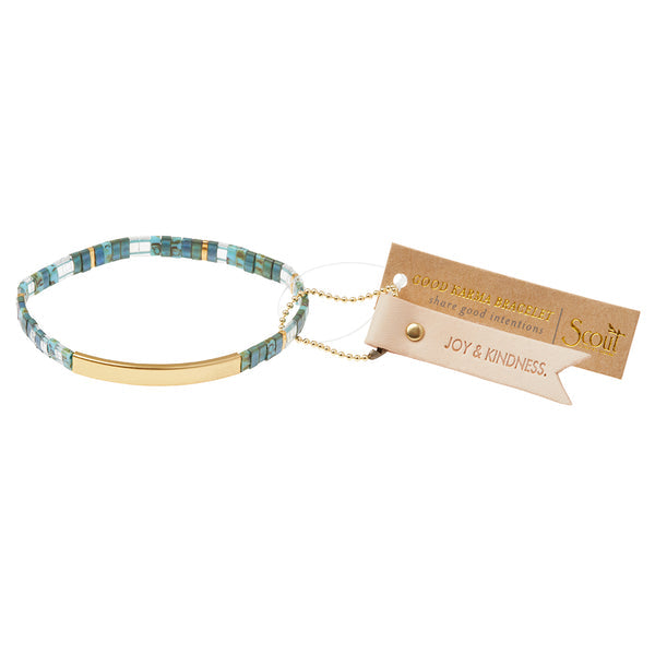 The Miyuki Joy and Kindness Marine Gold bracelet, by Scout Jewelry, showcases alternating light and dark blue square beads with a 14K gold-dipped bar accent. It comes with a brown tag labeled "GOOD KARMA BRACELET" and a beige tag that reads "JOY & KINDNESS," promoting messages of peace and positivity.