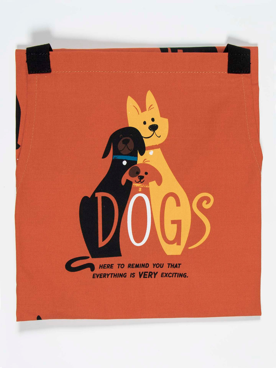 The Blue Q Dogs Apron features a whimsical design of three cartoon dogs—a yellow one, a black one, and a brown one—sitting together on an orange fabric with black velcro strips at the top. The text says, “DOGS here to remind you that everything is VERY exciting.” This charming scene is printed on heavy cotton twill for added durability.