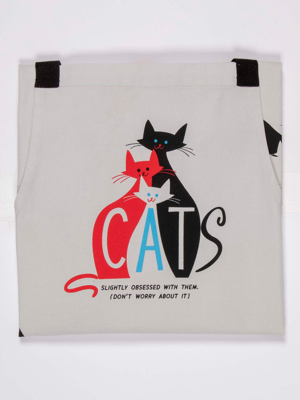 The Blue Q Cats Apron features an illustration of three cats in varying colors spelling out "CATS," accompanied by the phrase, "Slightly obsessed with them. (Don't worry about it)." The design is presented on sturdy cotton twill fabric with a light beige background.