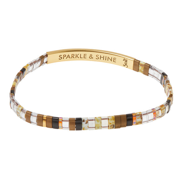 The Miyuki Sparkle and Shine Topaz Gold bracelet from Scout Jewelry features an elegant design with small square beads in shades of brown, clear, and gold. It is complemented by a 14K gold-dipped bar engraved with the message "SPARKLE & SHINE," inspired by Japanese glass Miyuki bracelets to offer an empowering message to its wearer.