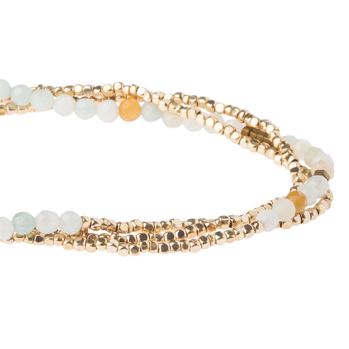 A detailed view of the Amazonite Gold Delicate Wrap by Scout Jewelry displays light-colored semi-precious gemstone beads interspersed with small gold accents. The stones blend in shades of white, pale blue, and orange, resulting in an elegant and delicate design as the bracelet coils into a circular shape.