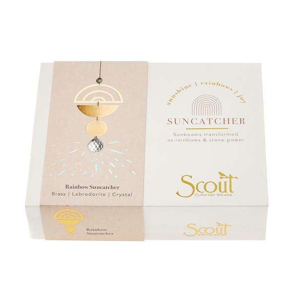 A packaging of a "Rainbow Labradorite Suncatcher" from Scout Jewelry. The box features a suncatcher decorated with brass, labradorite, and crystal. The text reads, "Sunbeams transformed as rainbows & stone power." The box combines beige and white colors with gold accents.