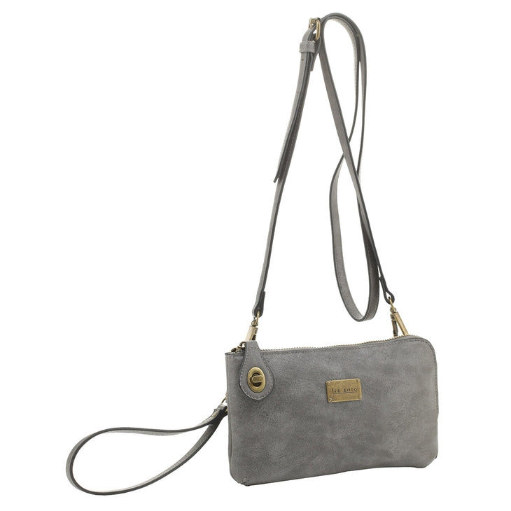 The Tina Cell Bag - Grey by Liz Soto Handbags is a stylish crossbody bag made of soft suede material, featuring a gold-tone zipper and logo plate. This small handbag includes an adjustable strap, a small tag with button decoration, and a compact design perfect for carrying essentials.