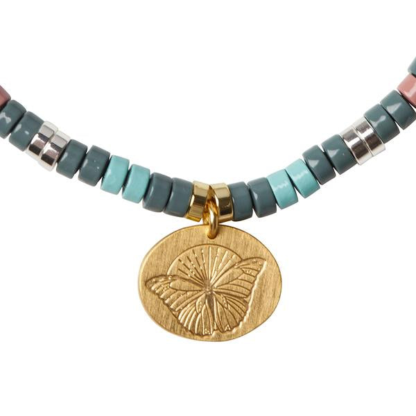 A close-up of the Intention Charm African Turq/Gold necklace by Scout Jewelry, showcasing various shades of turquoise, grey, and rose semi-precious stones. In the center hangs a round, gold-colored pendant with an embossed butterfly design that serves as a positive intention charm.