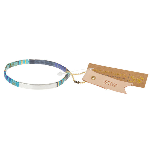 The Miyuki Brave Cobalt Silver bracelet by Scout Jewelry features small rectangular Japanese glass Miyuki beads in shades of blue, green, purple, and white. Attached is a brown tag with empowering messages and a beige tag inscribed with "BRAVE." A small gold chain connects the tags to the bracelet, creating a charming Scout Charm.