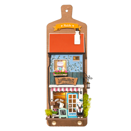 The DIY Miniature Wall Hanging Kit: Aroma Toast Lab by Hands Craft features a charming miniature cafe model on a cutting board. The blue building with a red roof has real lighting, doubles as decor and key holder, and includes a small figure under the awning and greenery for added life.