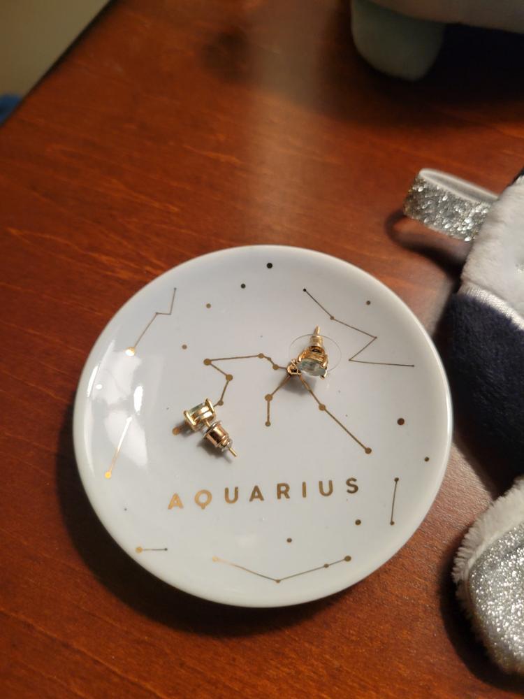 The LuckyFeather Dish Aquarius is a small white trinket dish with gold constellation accents and "AQUARIUS" in gold, shown holding gold stud earrings on a wooden surface—a stylish jewelry dish and thoughtful gift for her.