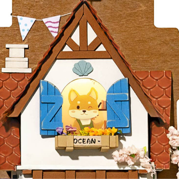 The DIY Miniature Wall Hanging Kit Island Dream Villa by Hands Craft makes a perfect gift idea, featuring a whimsical illustration of a dog in a fairy-tale house. The design includes blue shutters, a wooden roof, and decorative flags. The window frame is adorned with colorful flowers and the name "OCEAN" inscribed below, capturing the imagination of crafters and collectors alike.