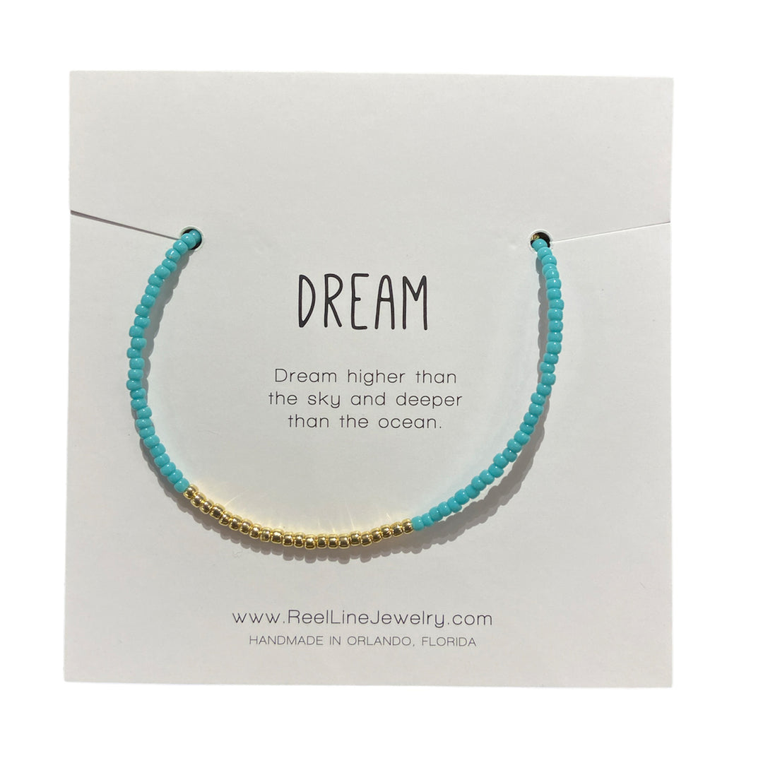 The "Dream - Turquoise Gold" beaded bracelet by Reel Line Jewelry elegantly features turquoise and gold beads on a card labeled "DREAM," inscribed with, "Dream higher than the sky and deeper than the ocean." This thin glass bracelet boasts adjustable links for a perfect fit. Handcrafted in Orlando, Florida.