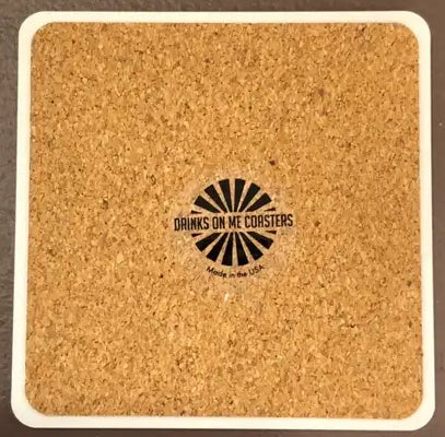 A square, light-brown "Lyrics Coaster" with a white border features the text "Drinks On Me" and "Made in the USA" printed in the center. The text is surrounded by a radiating black sunburst design, and the sturdy cork backing ensures durability and prevents slipping.