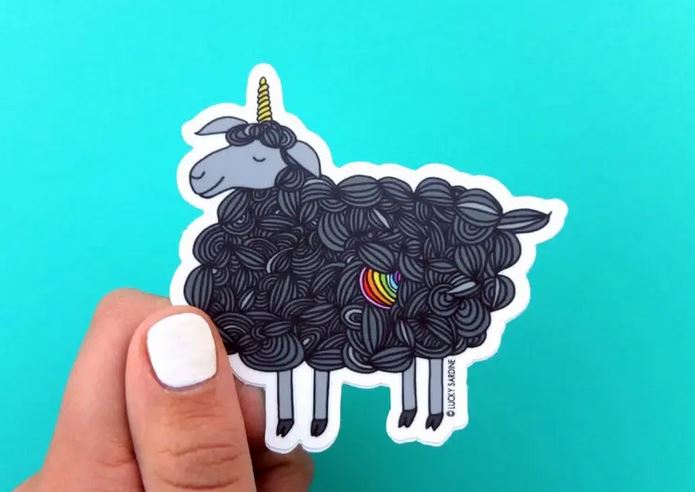 A hand with white-painted nails holds the Lucky Sardine's Black Sheep Unicorn Sticker, a scratch-resistant vinyl decal depicting a black sheep with a unicorn horn. The curly black wool of the sheep is adorned with a small rainbow heart, set against a solid teal background that highlights the vibrant details of this resilient sticker.