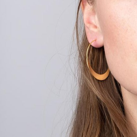 Close-up of a woman's ear adorned with a large Crescent Hoop Gold earring from Scout Jewelry. The thin, hypoallergenic, crescent-shaped piece complements her long brown hair, partially visible against the plain, light-colored background.