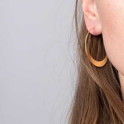 Close-up of a person’s ear adorned with a large silver Crescent Hoop earring by Scout Jewelry. The person's light brown hair is partially visible, cascading down their shoulder. This hypoallergenic piece blends elegance with comfort, ensuring no irritation for sensitive skin. The background is a soft gray.