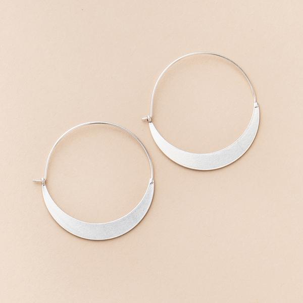 A pair of Scout Jewelry's Crescent Hoop Silver earrings, featuring a crescent-shaped design at the bottom, lying flat on a beige surface. Hypoallergenic and nickel-free, these sterling silver earrings boast a minimalist and modern appearance.