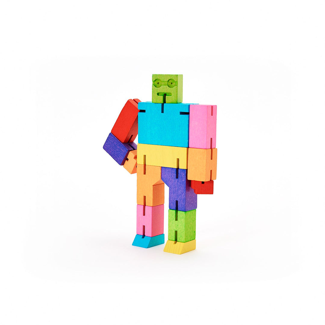 The Cubebot Multi Small from Areaware is a vibrant wooden toy robot that evokes the charm of Japanese Shinto puzzles. Its hinged limbs, crafted from blocks in green, blue, pink, orange, purple, and red hues, allow it to stand upright or bend at the joints. This playful figure boasts a square head with a minimalist face featuring two eyes and a mouth. One of its arms is bent at the elbow.