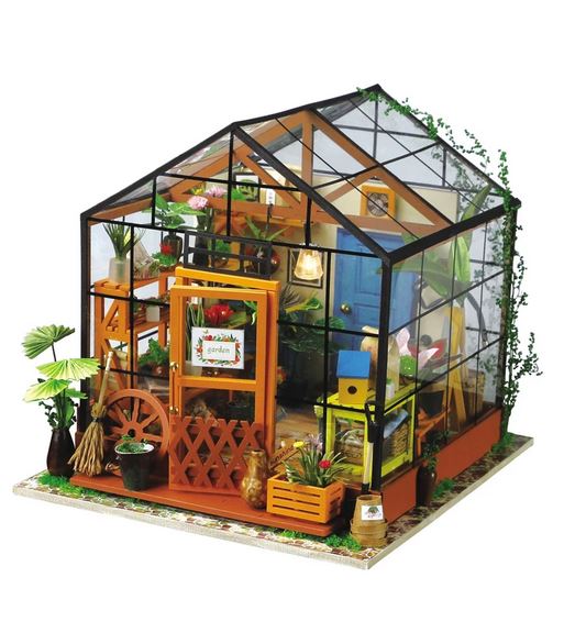 This image shows a small, detailed model of Cathy’s Flower House DIY Miniature Kit from Hands Craft, featuring transparent walls, a pitched roof, and wooden framing. Inside the 3D dollhouse puzzle, various miniature plants, gardening tools, and shelves are meticulously arranged. A sign on the door reads "garden," while the surrounding base displays more plants and greenery.