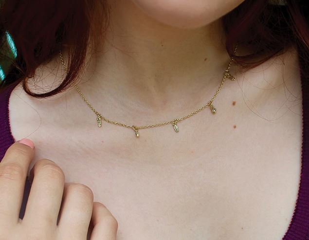 A person showcases the elegant Dangle Necklace Feather by LuckyFeather, an adjustable piece adorned with small vertical charms and plated in 14k gold. The blurred background accentuates the exquisite jewelry against a purple top.