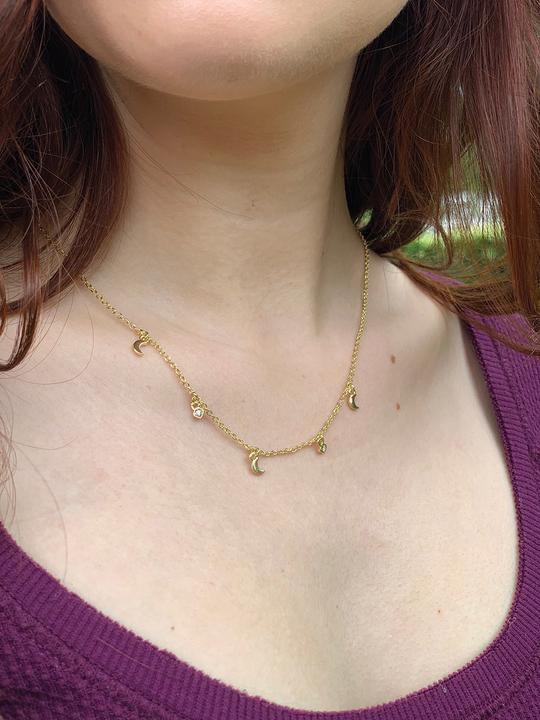 A close-up of a person wearing the LuckyFeather 'Dangle Necklace Moon,' an adjustable 14k gold plated chain adorned with small crescent moon and star charms. The person is dressed in a deep purple top, set against slightly blurred greenery.