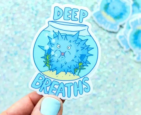 A hand adorned with light blue nail polish holds the Lucky Sardine "Deep Breaths Fish Bowl Sticker," featuring a surprised pufferfish inside a fishbowl. The words "Deep Breaths" are elegantly inscribed above and below the bowl. The backdrop is a sparkling, light blue surface.