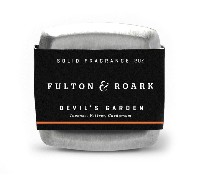 A silver tin of Fulton & Roark's solid fragrance, "Devils Garden," showcases a blend of rich incense and cardamom notes. It is elegantly wrapped in a sleek black and orange paper band and prominently displays the weight as .2 oz.