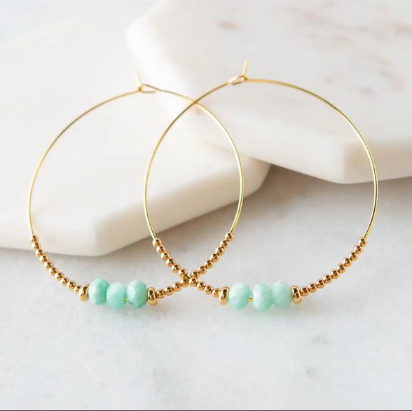 The Amazonite Lily Earrings by Lenny & Eva feature a delicate design, adorned with small turquoise beads and golden accents. This exquisite piece of handcrafted jewelry is elegantly displayed on a marble surface.