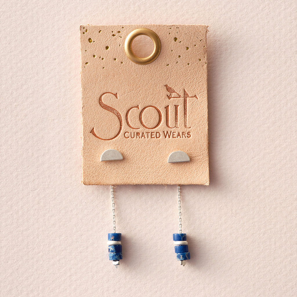 The Scout Jewelry "Meteor Thread Lapis Silver" earrings, hypoallergenic and free of nickel and cadmium, feature blue beads and silver chains elegantly displayed on a rectangular leather tag. A round gold-colored hole adorns the top.