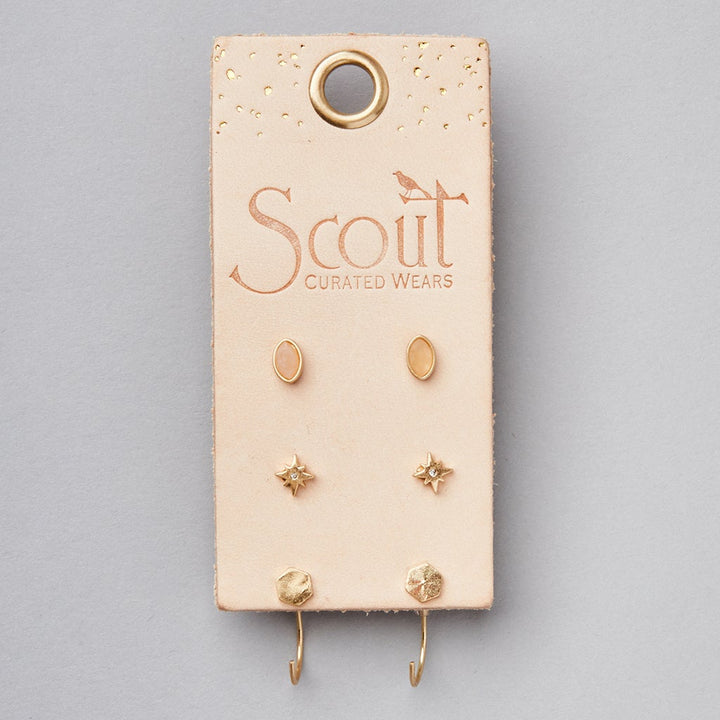 A light gray display featuring a beige earring holder from "Scout Jewelry" showcases the Gabby Stud Trio Gold, which includes three pairs of small, gold hypoallergenic stud earrings in oval, star, and hexagonal shapes.