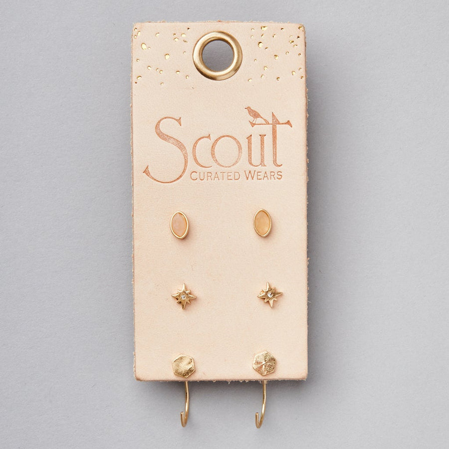 A light gray display featuring a beige earring holder from "Scout Jewelry" showcases the Gabby Stud Trio Gold, which includes three pairs of small, gold hypoallergenic stud earrings in oval, star, and hexagonal shapes.