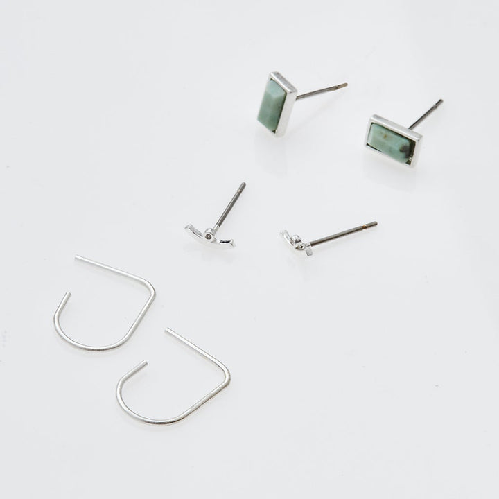 Three pairs of earrings from the Courtney Stud Trio Silver by Scout Jewelry, including silver U-shaped hoop earrings, brass rectangular green gemstone stud earrings, and tiny bow-shaped hypoallergenic stud earrings, are laid out on a white background.