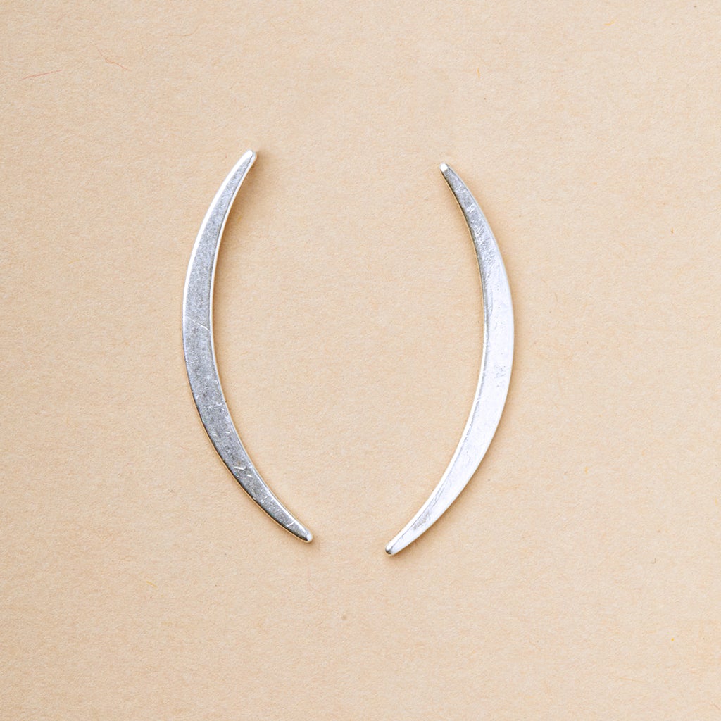 On a light beige background, the Gibbous Slice Stud Silver earrings by Scout Jewelry display two symmetrical curved metal pieces like crescents or elongated parentheses. Part of the Refined Earring Collection, they are crafted in exquisite 18k gold vermeil and 925 sterling silver.