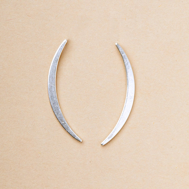 On a light beige background, the Gibbous Slice Stud Silver earrings by Scout Jewelry display two symmetrical curved metal pieces like crescents or elongated parentheses. Part of the Refined Earring Collection, they are crafted in exquisite 18k gold vermeil and 925 sterling silver.