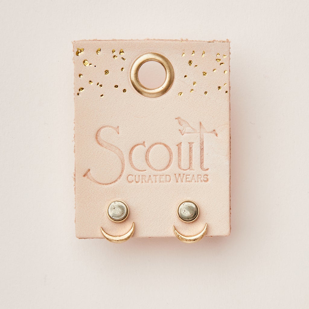 Image of Moon Phase Pyrite/Gold earrings displayed on a beige card with "Scout Jewelry" printed on it. The hypoallergenic stud earrings feature a clear, semi-precious stone set in a circular metal frame with a small crescent moon shape below. The card has a hole at the top and gold accents.