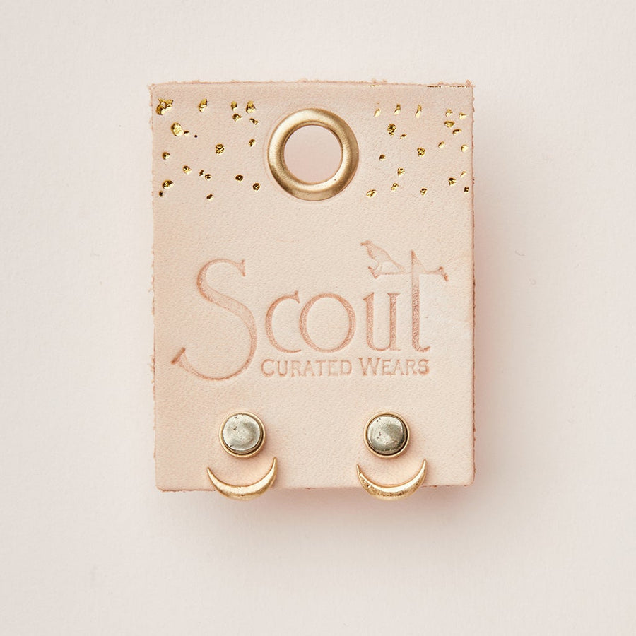 Image of Moon Phase Pyrite/Gold earrings displayed on a beige card with "Scout Jewelry" printed on it. The hypoallergenic stud earrings feature a clear, semi-precious stone set in a circular metal frame with a small crescent moon shape below. The card has a hole at the top and gold accents.