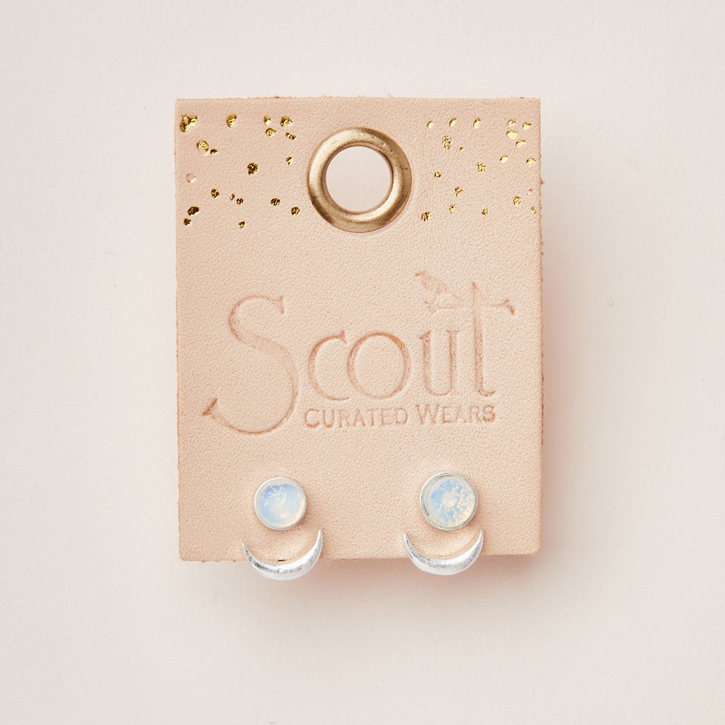 A pair of semi-precious Moon Phase Amazonite/Gold earrings is displayed on a beige leather card embossed with "Scout Jewelry." The earrings feature a small round amazonite at the top with an adjustable brass crescent ear jacket underneath. The card is adorned with gold speckles and has a loop hole at the top.
