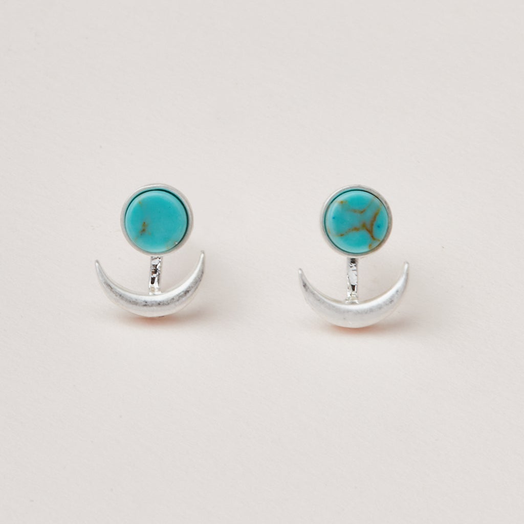 Introducing the Moon Phase Turquoise/Silver earrings from Scout Jewelry: These hypoallergenic earrings feature a round turquoise stone on top, complemented by a silver crescent moon shape suspended below. Set against a plain and light-colored background, their simplicity and elegance are beautifully showcased.