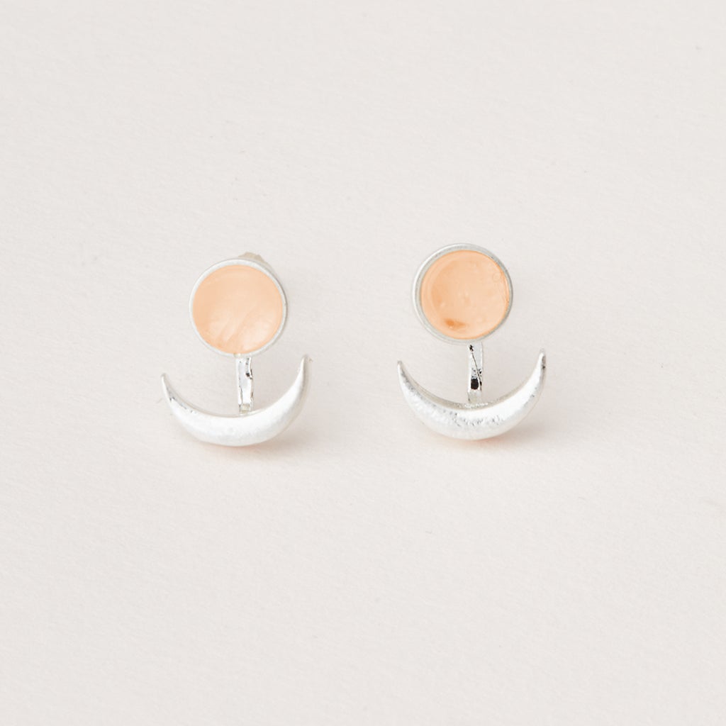 A pair of minimalist earrings from Scout Jewelry, named the Moon Phase Sunstone/Silver, features adjustable brass crescent moons beneath small circular semi-precious peach-colored stones. The earrings are displayed on a plain white background.