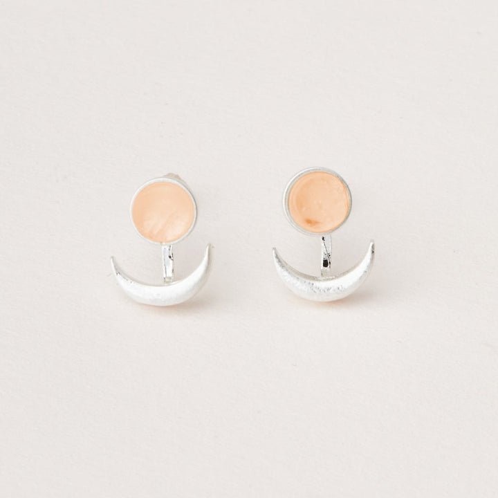 A pair of minimalist earrings from Scout Jewelry, named the Moon Phase Sunstone/Silver, features adjustable brass crescent moons beneath small circular semi-precious peach-colored stones. The earrings are displayed on a plain white background.