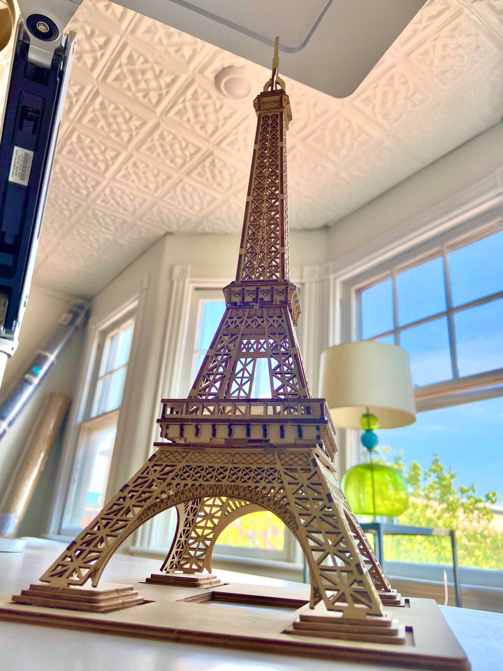 A detailed Hands Craft 3D Wooden Puzzle: Eiffel Tower sits on a white table in a bright room with large windows. The room, reflecting a modern vintage style, has white walls, an ornate patterned ceiling, and a green table lamp by the window. Sunlight illuminates the space beautifully.