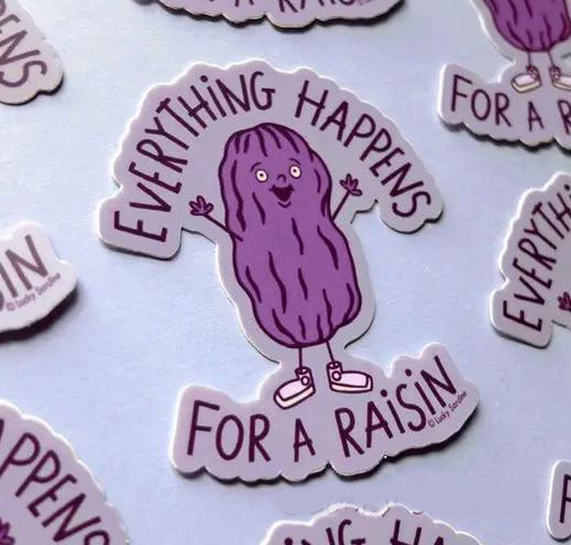 A whimsical, waterproof sticker featuring a cheerful, anthropomorphic raisin with small arms and legs, donning sneakers and raising its hands. The text around it reads "Everything Happens For a Raisin." Stickers identical to this one are scattered in the background. This is the "Everything Happens For a Raisin Sticker" from Lucky Sardine, made from durable vinyl.