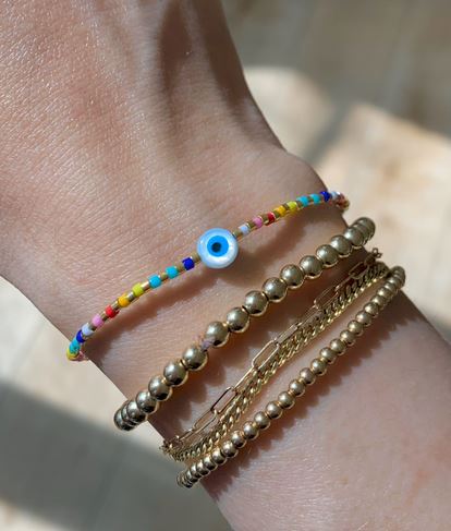 A wrist adorned with Kindred Row's Good Vibes Eye Stacking Bracelet showcases a colorful design featuring natural gemstones and a blue eye charm, elegantly paired with multiple gold bead and chain bracelets. The background is softly blurred.