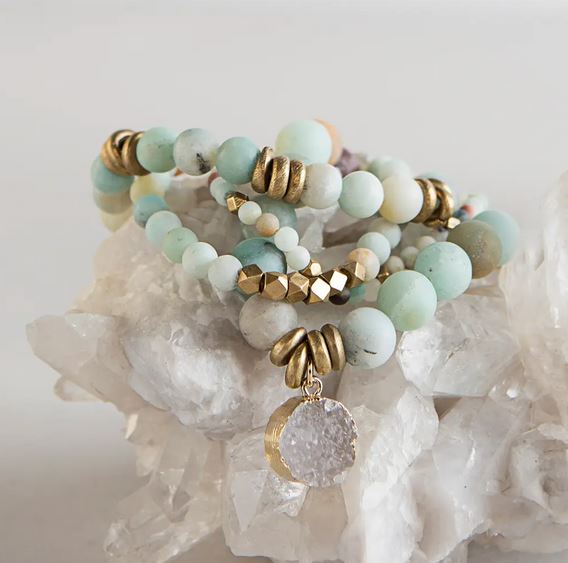 A beautiful set of Lenny & Eva's Amazonite Gemstone Bracelets, featuring light blue and white marbled beads, brass accents, and a gold-framed amulet with a rough crystal pendant. The stylish natural semi-precious 4mm gemstones are elegantly displayed on a cluster of white quartz crystals and stretch to fit most wrists.