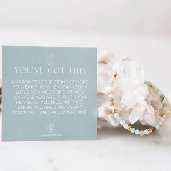 Displayed next to the Amazonite Gemstone Bracelet from Lenny & Eva is a card with an inspirational message. The card reads: "You've got this. Amazonite is the stone of hope. So wear this when you need a little reminder of just how capable you are." Adorned with light blue and gold beads along with brass accents, the bracelet, measuring 4mm and designed to fit most wrists, drapes elegantly over a white crystal cluster.