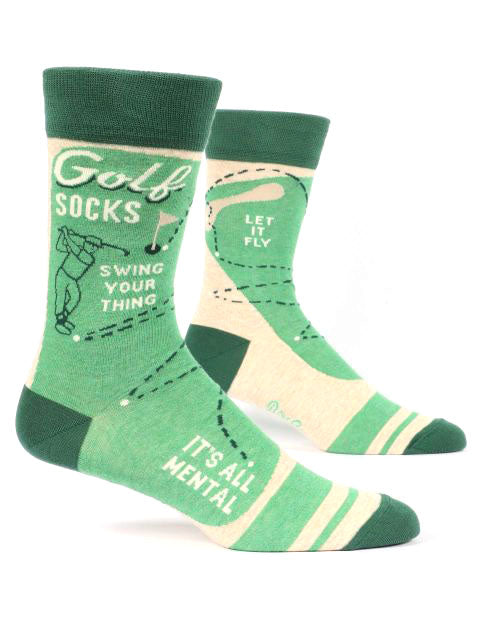 The Blue Q Golf Socks are a pair of green and beige cotton socks featuring various golf-related designs and text. One sock reads "Golf SOCKS," "Swing Your Thing," and "It’s All Mental," while the other reads "Let It Fly." The design includes illustrations of a golfer and a golf course. They fit men's shoe sizes 7-12 perfectly.