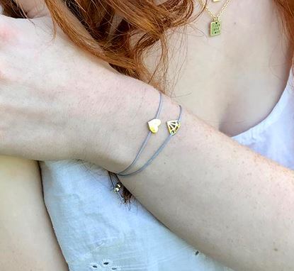 A person celebrates their birthday wearing LuckyFeather's Love Birthday bracelet, elegantly designed with delicate heart and triangle charms. They are dressed in a white, sleeveless top, with light brown curly hair cascading down as they also wear another necklace featuring a stunning gold pendant.